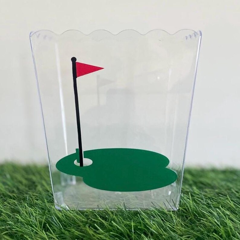 May include: Clear plastic container with a green golf course and a red flag on a black pole. The container has a scalloped edge.