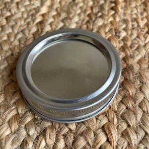 May include: A silver metal lid for a jar or container. The lid has a smooth, shiny surface and a slightly raised rim.