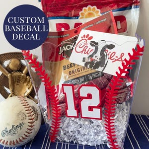 May include: A clear baseball-themed gift basket with red stitching details. The basket contains beef jerky, a gift card, and a baseball. A white number 12 is visible on the front. The text "CUSTOM BASEBALL DECAL" is on a blue circle.