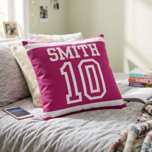 May include: A square magenta throw pillow featuring the white text SMITH and the number 10 in a bold athletic font. The design includes a horizontal white stripe across the top and bottom, mimicking a sports jersey aesthetic on the fabric.