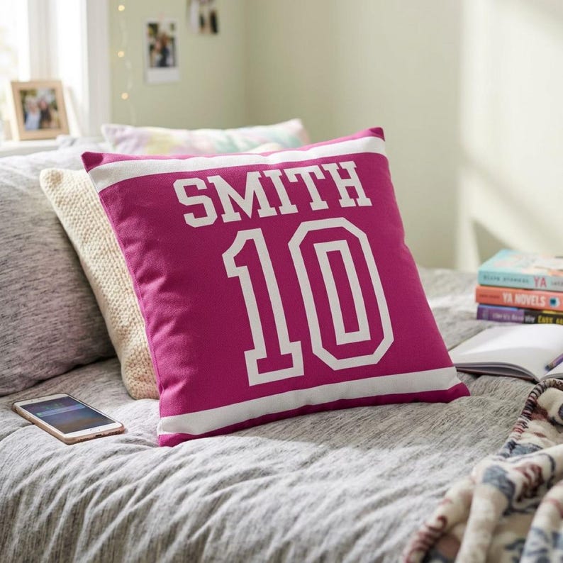 May include: A square magenta throw pillow featuring the white text SMITH and the number 10 in a bold athletic font. The design includes a horizontal white stripe across the top and bottom, mimicking a sports jersey aesthetic on the fabric.