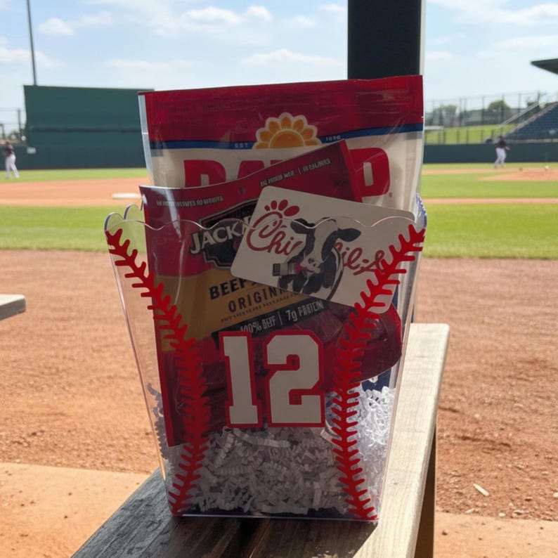 May include: A clear plastic baseball-themed gift basket filled with snacks and a gift card. The basket has red baseball stitching and the number 12. Inside are bags of beef jerky and a gift card with a cow logo.