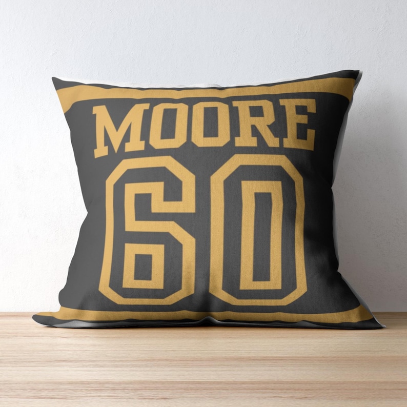 May include: A square throw pillow featuring a dark gray background with a gold border. The text MOORE is printed in large gold block letters above the number 60, which is also displayed in large gold block lettering in a sports jersey style.