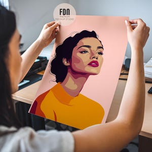 May include: A woman holds up a poster featuring a colorful illustration of a woman with dark hair and a yellow shirt. The background is a light pink color.