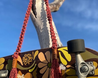 Könnte beinhalten: Ein Skateboard mit braunem Blumendesign und dem Text "GRL SWRL" wird gezeigt. Eine rostfarbene Makramee-Tasche mit Lederakzenten hängt an dem Board. Der Hintergrund zeigt einen blauen Himmel und einen verwitterten Ast.