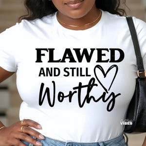 Puede incluir: Camiseta blanca con el texto negro "FLAWED AND STILL Worthy" y un corazón. La camiseta es de cuello redondo.