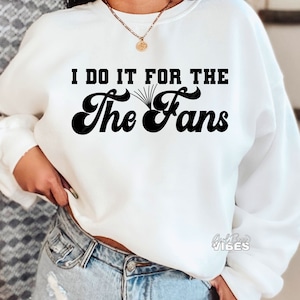 May include: White sweatshirt with black text that reads "I do it for the The Fans" in a retro font.