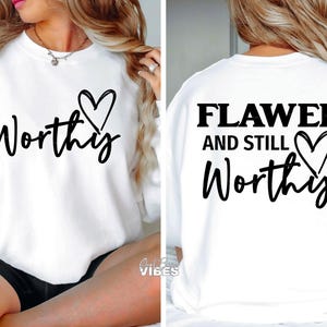 Op de afbeelding: Wit sweatshirt met het woord "Worthy" in zwart script en een zwarte hartafbeelding. De achterkant van het sweatshirt heeft de tekst "Flawed and Still Worthy" in het zwart. Een casual, comfortabele stijl.