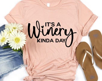Può includere: Una maglietta rosa con il testo "It's a Winery Kinda Day" in corsivo nero.