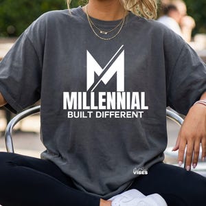 Könnte beinhalten: Dunkelgraues T-Shirt mit dem weißen Schriftzug "MILLENNIAL BUILT DIFFERENT" und einem weißen "M"-Grafik. Das T-Shirt hat eine lockere Passform und kurze Ärmel.