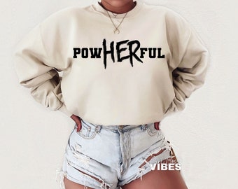 Könnte beinhalten: Ein beiger Pullover mit dem Aufdruck "POWHERFUL" in Schwarz. Der Pullover wird von einer Person mit Jeans-Shorts getragen.
