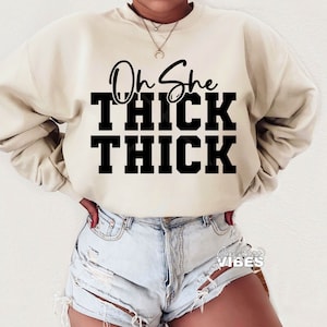 May include: A cream-colored sweatshirt with the text "Oh She Thick Thick" printed in black.