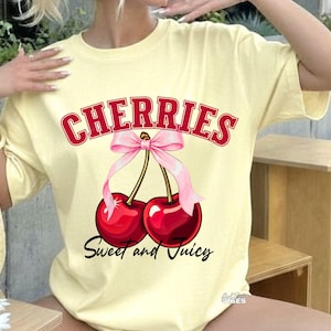 Op de afbeelding: Een lichtgeel T-shirt met het woord "CHERRIES" in rode blokletters met een witte omlijning. Daaronder een afbeelding van twee rode kersen die met een roze strik zijn vastgebonden. De tekst "Sweet and Juicy" staat onder de kersen.