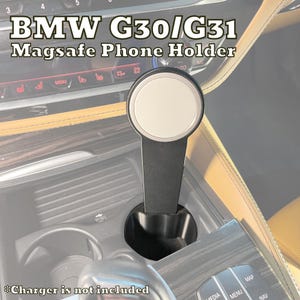 May include: A black MagSafe phone holder for BMW G30/G31 models. The holder features a circular magnetic charging pad with a silver border and a black stem that fits into a car's cup holder. The text "BMW G30/G31 Magsafe Phone Holder" is visible.