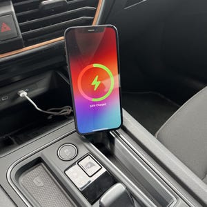 Puede incluir: Un teléfono inteligente negro cargándose en un coche. La pantalla muestra un icono de carga con un rayo verde y el texto "54% Charged". El teléfono está conectado a un cable de carga blanco.