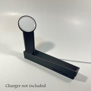 Könnte beinhalten: Schwarzer, abgewinkelter Ladeständer für eine Smartwatch mit einem kreisförmigen Ladepad oben. Der Ständer hat eine Ablage an der Basis und der Text "TheroParts" ist auf der Seite aufgedruckt. Der Text "Ladegerät nicht enthalten" steht unten.