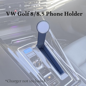 Peut inclure: Un support de téléphone noir pour véhicule VW Golf 8/8.5, doté d'un support vertical fin avec une tête de fixation magnétique circulaire. L'accessoire est conçu pour s'insérer dans la console centrale près du levier de vitesse. Texte : VW Golf 8/8.5 Phone Holder.