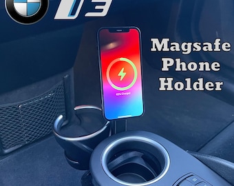 May include: A black MagSafe phone holder with a charging iPhone. The phone displays a charging symbol. The holder is mounted in a car's cup holder. The text "Magsafe Phone Holder" is visible.