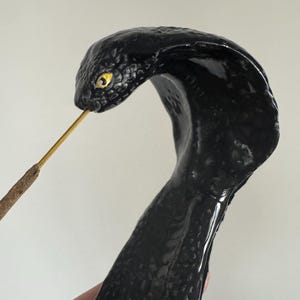 Op de afbeelding: Een zwarte cobra wierookhouder met een glanzende afwerking. De kop van de slang is omhoog, met een gele oog en een gat voor een wierookstokje. De houder is ontworpen om op een cobra slang te lijken.