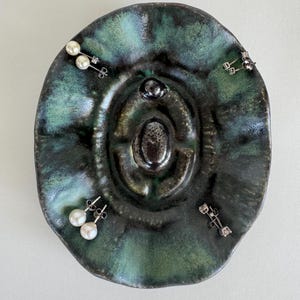 May include: A green ceramic dish with a raised center, designed to hold jewelry. The dish has three pairs of earrings on it, including pearl and diamond earrings.