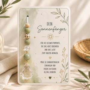 May include: A suncatcher ornament attached to a rectangular card featuring the German text 'DEIN Sonnenfänger' and a poem. The ornament consists of a white cord strung with wooden, sage green, patterned, and clear beads, ending in a faceted crystal drop.