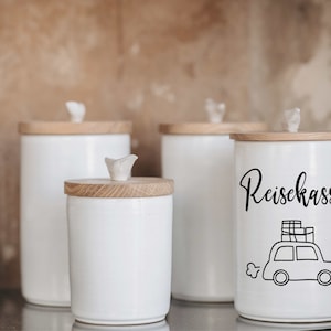 Może przedstawiać: Cztery białe ceramiczne pojemniki z drewnianymi pokrywkami. Pojemnik po prawej stronie ma napis "Reisekasse". Pojemnik ma czarno-białą ilustrację samochodu z bagażem na dachu.