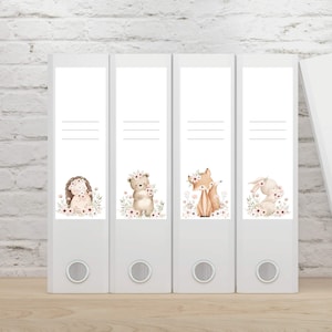 Könnte beinhalten: Vier weiße Ordner mit dekorativen Etiketten mit Illustrationen von Waldtieren. Die Etiketten zeigen einen Igel, einen Bären, einen Fuchs und ein Kaninchen, jedes umgeben von floralen Akzenten. Die Ordner sind auf einer hellen Holzoberfläche angeordnet.