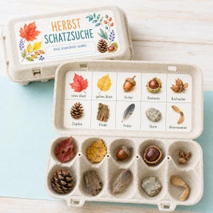 Könnte beinhalten: Ein hellgrauer Eierkarton aus Pappe für eine herbstliche Schatzsuche. Der Deckel trägt den Text HERBST SCHATZSUCHE sowie Illustrationen von Blättern und Nüssen. Zehn Fächer enthalten passende Naturmaterialien wie Blätter und Steine.