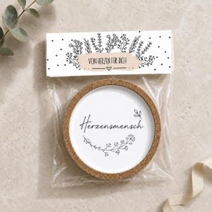 Puede incluir: Un posavasos redondo de corcho con un centro blanco que presenta el texto negro Herzensmensch y un gráfico floral. Está empaquetado en una funda de plástico transparente con una tarjeta de cabecera blanca con el texto VON HERZEN FÜR DICH.