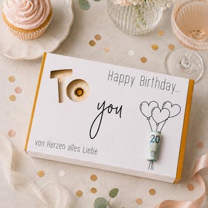 Pode incluir: Uma caixa de presente retangular branca com bordas douradas contendo o texto "Happy Birthday...", "To", "you" e "von Herzen alles Liebe". A caixa inclui um recorte circular e um gráfico de três balões com uma nota de 20 euros enrolada.