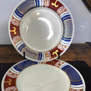 May include: A stack of white ceramic plates with a decorative border. The border features a pattern of blue, red, and gold geometric shapes and floral designs. The plates are stacked on a dark surface.