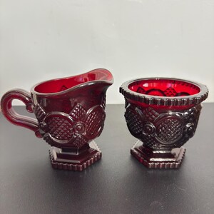 May include: A ruby red glass creamer and sugar bowl set. The creamer has a handle and spout, while the sugar bowl is open. Both pieces feature a detailed floral and geometric pattern. The set is displayed on a dark surface.