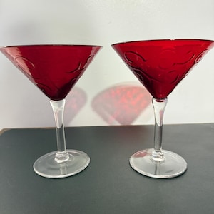 Könnte beinhalten: Zwei rubinrote Martinigläser mit klaren Stielen und Böden. Das rote Glas hat ein dekoratives geätztes Design. Die Gläser werden auf einer dunklen Oberfläche vor einem weißen Hintergrund präsentiert.