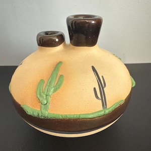 May include: A ceramic vase with a tan body, two dark brown spouts, and a dark brown base. The vase is decorated with green cacti and a green border. The vase is a decorative item.