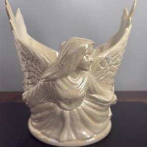 Pode incluir: Estatueta de anjo creme com asas abertas. O anjo de cerâmica tem uma expressão serena e usa uma túnica drapeada. As asas são detalhadas com texturas de penas. A estatueta tem um acabamento iridescente.