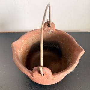 Peut inclure: Un petit pot en fonte brun rougeâtre avec une poignée en métal incurvée. Le pot a une forme arrondie avec un bord légèrement évasé et un petit bec de chaque côté. La poignée est fixée au pot par une boucle simple.