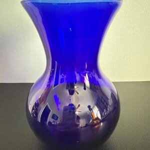 May include: A cobalt blue glass vase with a wide, rounded base and a flared top. The vase has a smooth, glossy finish and reflects the surrounding environment. The vase is a decorative item.