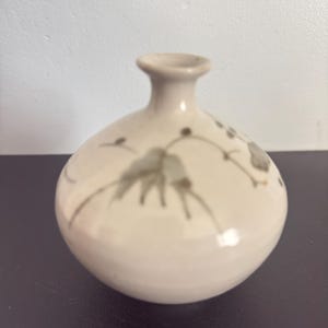 May include: A small, cream-coloured ceramic vase with a rounded body and a narrow neck. The vase features a delicate, hand-painted floral design in shades of grey. The vase is on a dark surface.