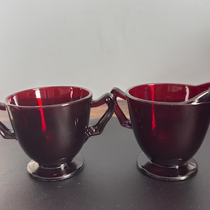 以下が含まれることがあります： ルビーレッドのガラス製シュガーボウルとクリーマーのセット。シュガーボウルには2つのハンドルと丸みを帯びたベースがあります。クリーマーには注ぎ口と似たようなベースがあります。どちらのアイテムも濃い赤色のガラス製です。