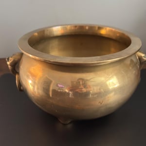 May include: A golden-colored metal bowl with a wide rim and three small feet. The bowl has two decorative handles on either side. The interior of the bowl is visible, and the surface reflects the surrounding environment.