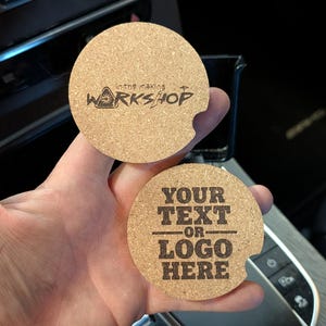 May include: Two round cork car coasters. One coaster has the text "in the making WORKSHOP" in black. The other coaster has the text "YOUR TEXT OR LOGO HERE" in black.