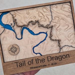 May include: Wooden framed map of the Tail of the Dragon, US 129, featuring a detailed topographic design with a blue river and a compass rose. The text "Tail of the Dragon" and "318 curves in 11 miles" are engraved on the frame.