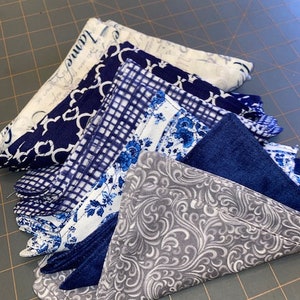 Peut inclure: Un empilement de cinq carrés de tissu dans différents motifs bleus et blancs, y compris un carré gris avec un motif floral blanc. Les carrés de tissu sont pliés et disposés en pile.