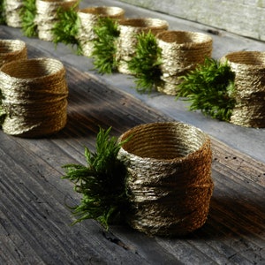 Könnte beinhalten: Goldene und braune Burlap-Serviettenringe mit grünen Zweigen aus Grünzeug, die an jedem Ring befestigt sind. Die Ringe sind in einer Reihe auf einer rustikalen Holzoberfläche angeordnet.