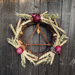 May include: A circular wreath crafted from woven brown twigs, decorated with dried wheat, burgundy flowers, and small stones. A wooden triangle is at the centre, with a small crystal pendant suspended from it. The wreath is set against a weathered wooden backdrop.