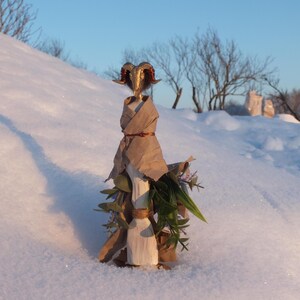 May include: A handcrafted sculpture of a ram's head with golden horns, adorned with paper and greenery. The sculpture is wrapped in brown paper and decorated with leaves and small purple flowers. The artwork is set against a snowy backdrop.