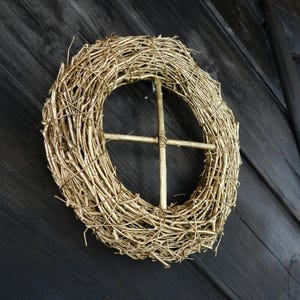 May include: A gold-painted twig wreath with a decorative metal cross in the centre. The wreath is hanging on a dark wooden wall.