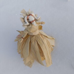 May include: A handmade doll crafted from dried corn husks, featuring a skirt-like base and a detailed head with horns. The doll's arms are tied with thin copper wire, and it is set against a snowy background.