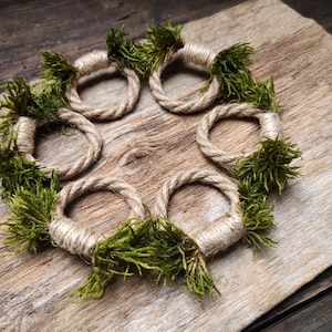 Pode incluir: Um conjunto de seis argolas de guardanapo de corda de juta natural decoradas com musgo verde. As argolas estão dispostas em círculo sobre uma superfície de madeira rústica.