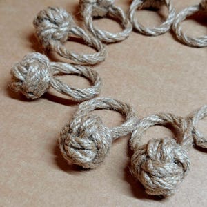 May include: Close-up of several handmade rope napkin rings. Each ring features a decorative knot and a circular loop, all made from natural-coloured rope. The rings are arranged on a light brown surface, suggesting a rustic or nautical theme.